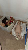 Large box with four assorted plush toys including teddy bears and a snowman, next to a cardboard box containing the 21-inch antique stockinette doll wrapped in tissue paper laying on carpet.