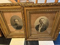 Full view of both framed vintage portraits leaning against a wall on a tiled floor.