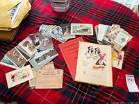 Wide view of vintage postcards and ephemera spread on a red and black plaid cloth, showing various old postcards, including colored portrait cards, booklets, and printed papers.