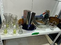 Various vintage stemware glasses with greenish-yellow stems alongside carnival glass pieces and barware items arranged on white shelf.