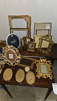 Photo showing a variety of approximately 18 wood and metal picture frames arranged on two surfaces, including ornate gold-toned carved frames, oval and rectangular shapes, some with velvet or fabric backing inside.