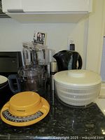 Overall lot view showing all items: food processor with attachments, kitchen scale, electric kettle, and salad spinner on a kitchen countertop.
