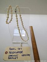 Full shot of the freshwater pearl necklace, bracelet, and single earring displayed on a clear acrylic stand next to a ruler.