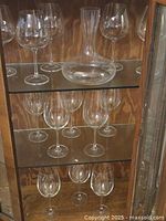 Wooden curio cabinet with glass shelves and glass-panel door, containing clear glass stemware and a clear glass decanter.