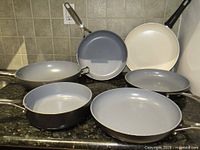Six frying pans and saucepans arranged on countertop showing mixed colors and sizes, mainly gray and off-white ceramic interiors with stainless steel handles.