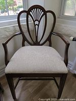 Front view showing wooden frame arm chair with beige fabric upholstered seat featuring subtle swirl pattern and carved openwork shield backrest with fan motif.