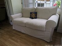 Front view of the beige/cream two-seat sofa with rounded arms and skirted base, including a decorative pillow with sun motif.