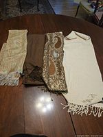 Three scarves laid out on a table. From left to right: beige paisley patterned scarf with fringe, solid brown scarf, brown paisley patterned scarf with fringe.