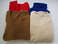 Four folded cashmere sweaters in red, blue, brown, and off-white colors, photographed from above.