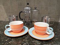 Photo showing two French presses, two stainless steel cups, and two orange swirl ceramic coffee cups with matching saucers on countertop