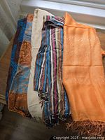 Four scarves/wraps folded and layered showing different color patterns and textures including orange jacquard, striped, blue patterned, and neutral tone