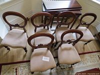 Set of six Victorian balloon back mahogany dining chairs with beige upholstered seats, arranged in two rows on hardwood floor and near a rug. Showcases the carved floral motif on back and turned legs with casters.