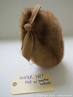 Side view of tan-colored fur winter hat with ponytail cutout and side fabric bow.