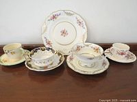 Four assorted vintage bone china and porcelain tea cup and saucer sets displayed on a wooden surface with one decorative floral plate behind them. Each cup and saucer set has distinct shapes, scalloped edges, and floral patterns with gold trim.