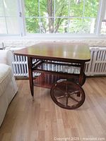 Front angled view of vintage wooden tea cart with foldable sides, removable glass tray on top, and large spoked wooden wheel showing wear.