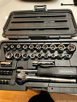 Opened case showing silver metal socket set including various sizes, a ratchet wrench, extension bar, arranged inside black molded plastic insert.