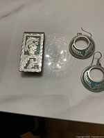 Two earrings and one money clip arranged on a white surface showing detailed embossing and turquoise inlay on earrings