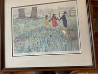 Framed art print showing three children walking through a field of yellow flowers with trees and a fence in the background. The print is matted and framed.