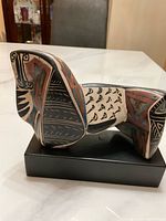 Side view showing geometric painted patterns and abstract animal form shape on ceramic sculpture mounted on black base.