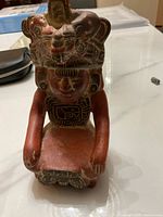 Front view of reddish-brown ceramic sculpture vase depicting a seated figure with animal headdress, showing detailed facial and body carvings