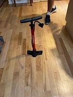 Manual bicycle pump standing upright on wood floor, showing red barrel, black handle, and foot base