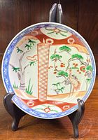 Full view of the vintage Imari porcelain bowl on black display stand showing detailed crane and foliage hand-painted artwork inside bowl bowl rim decorated with blue wave pattern