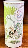 Cylindrical porcelain vase with scene of figure riding an animal under green leaves and tree, detailed hand painting and calligraphy above.