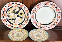 Front view of four antique transferware plates showcasing two larger orange and black patterned plates and two smaller blue and gold floral patterned plates.