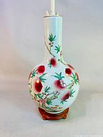 Full view of globular Famille Rose porcelain vase with peach motif on wooden stand and lamp hardware attached.