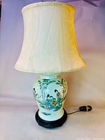 Full view of the table lamp showing the porcelain ginger jar body with hand-painted Chinese figure scenes, fabric lamp shade, dark carved wooden base, and electrical cord.