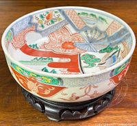 Japanese Imari porcelain bowl hand-painted with polychrome detail of trees, flowers, and a bird, resting on carved black wooden stand.