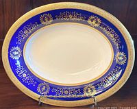 Full front view of the oval bowl showing the cobalt blue rim with detailed gold encrusted floral and ornamental designs surrounding the white interior.