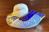 Porcelain duck paperweight with white, cobalt blue, and gold details. Shown from side angle on wooden surface.