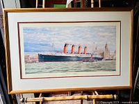 Framed print showing The Lusitania ship in the harbor with buildings in the background, gold-tone frame visible.