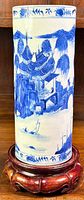 Full view of tall cylindrical blue and white porcelain vase on wooden stand, depicting continuous landscape scene.