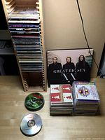 Wooden CD rack with a tall stack of CDs, several stacked CDs including karaoke and Christmas themed, and a vinyl album by Great Big Sea