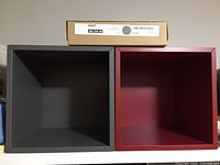 Two EKET IKEA square shelf units side by side, one dark gray and one dark red, with one new in box on top