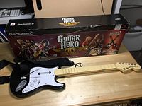 Black Guitar Hero guitar controller with beige fretboard and white pickguard lying in front of original boxed packaging for Playstation 2 version.