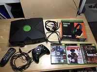 Photo showing original black Microsoft Xbox console, black wired controller, remote control, cables, Tritton Kama Xbox headset box, and three Xbox game cases (Madden NFL 07, ESPN NBA 2K5, Rainbow Six 3) on a wooden table.