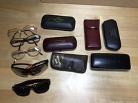 Top down photo of six pairs of eyeglasses and sunglasses with seven cases arranged on a table, showing brands and frame styles.