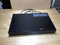 Top angled view showing the black Samsung Blu-Ray DVD player with power cord on a table.