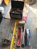 Full lot overview showing the black metal toolbox with assorted hand tools, a yellow spirit level, concrete mixer paddle, and other items laid out on the floor.