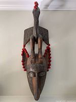 Front view of the large carved wooden Bambara tribal mask featuring a bird on top and red pom-poms on the sides.