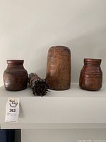 Front view showing three wooden storage pots of various shapes with a bundle of sticks placed in middle on a shelf.