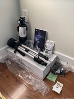 Telescope optical tube, tripod folded, manual, and accessories laid out in corner on floor