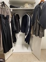Open white closet showing multiple garments including a black dress, fur coat, leather coat, blazers and garment bags