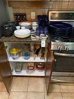 Photo of kitchen counter and cabinet open showing blue glass dishes, black bowls, white pasta bowls, mugs, coasters, and various kitchen tools.