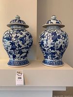 Front view of the two matching floral patterned blue and white porcelain urns with lids placed side by side on a white surface.
