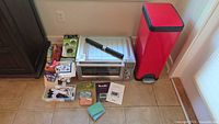 Front and top view of the Breville stainless steel toaster oven with the red garbage bin to its right and assorted kitchen accessories around it on a tiled floor.