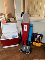 Coleman cooler, Under Armour lunch box, hiking sticks, folding chair in carry bag, blue mat, life jacket on wooden floor.